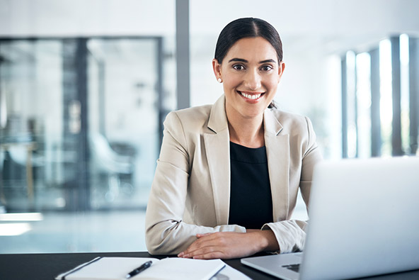 A young woman sitting at her office desk is smiling at the camera. She has brown hair and  eyes and is wearing a black shirt and beige coloured jacket.